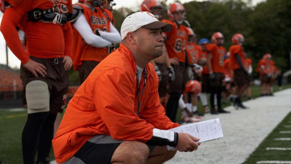 Coach squatting on the sidelines watching a game while wearing custom football warmups